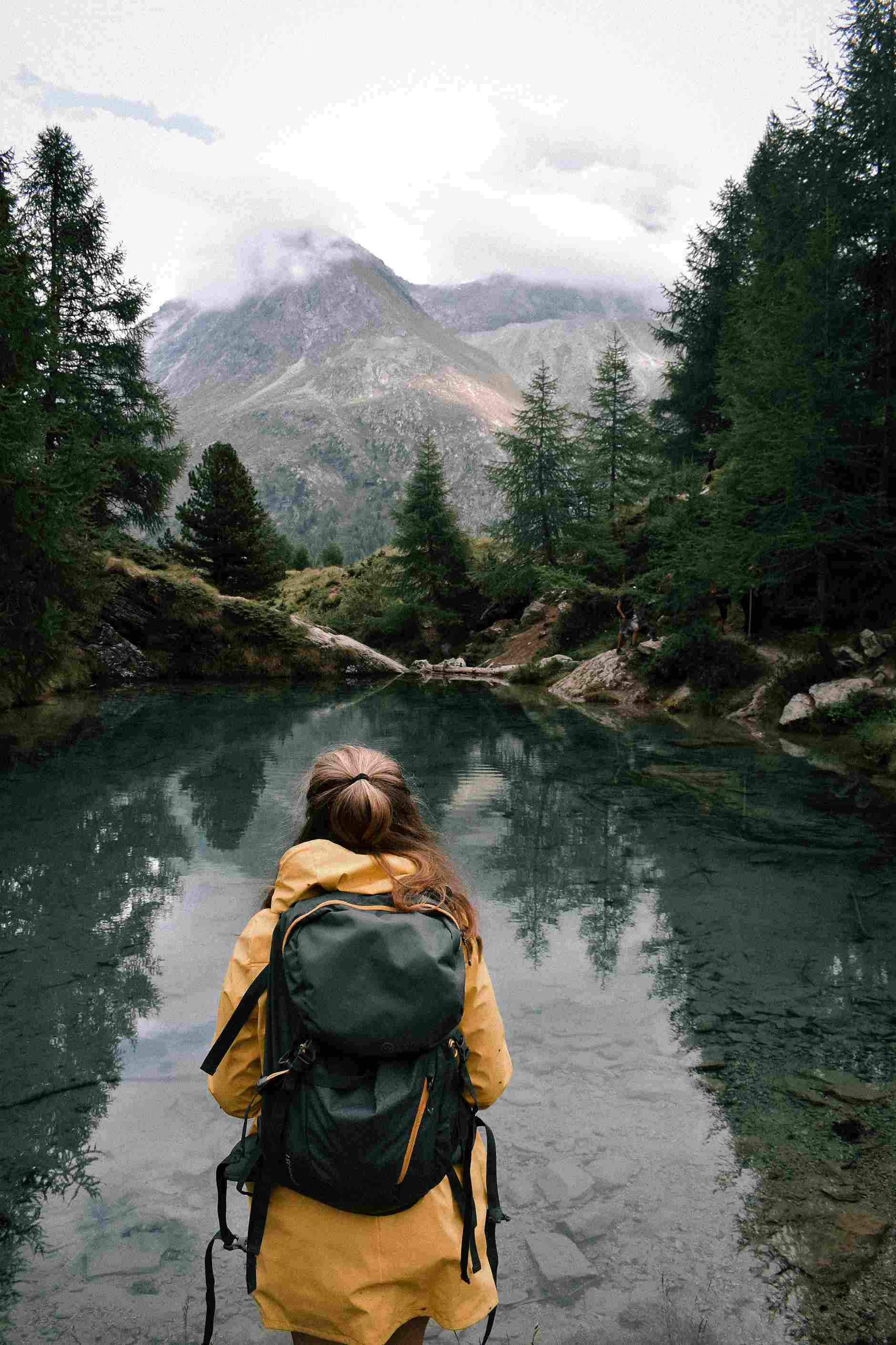 A lady in a yellow jacket, looking out over a green water with mountains in the background surrounded by 
             a beautiful landscape with green trees around the water.